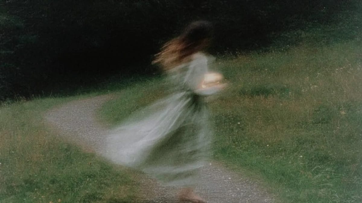 Soft-focus photo of a woman in a pale dress moving along a curving path in a green field, motion blur trailing her skirt.