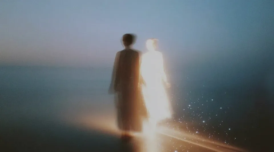 Soft-focus scene of a figure on a shoreline at dusk, one dark, one glowing white with sparkles, walking toward the horizon.